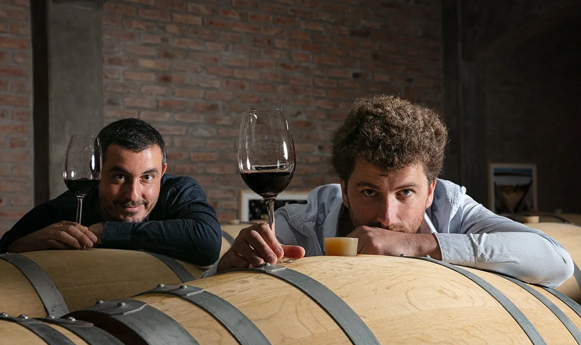 Quentin et Thibault, verre de vin en main, sur les barriques